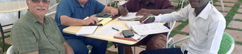 Eaar Oden, Bud Potter, Kiefa Moseti Ontiri and Eric Mogendi meeting at a table under a large white tent in Kenya, a close up of their table with their note pads, pens and hands.